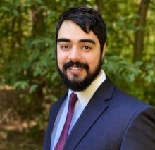 A man with a beard, dressed in a suit, against a backdrop of trees.